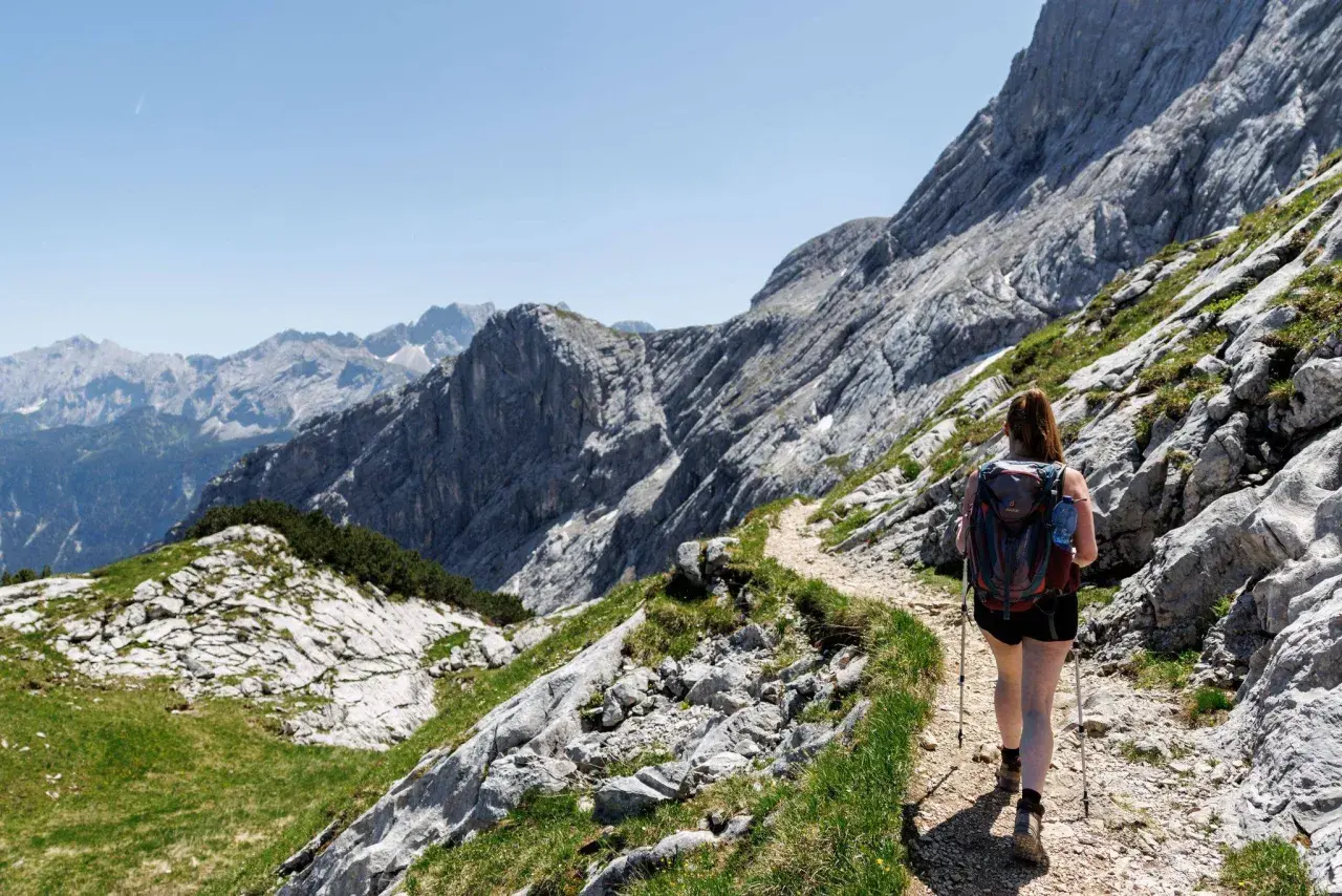 Frau mit Rucksack und Wanderstöcken genießt das Wandern in den Alpen auf einem steinigen Pfad mit Blick auf majestätische Berge.