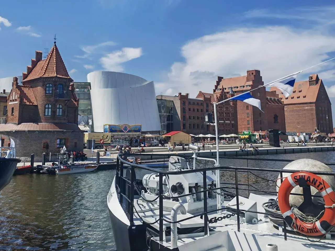Hafenansicht mit historischer Backsteinarchitektur und modernem Gebäude. Ein Schiff mit Rettungsring liegt im Vordergrund. Stralsund sehenswertes Panorama.