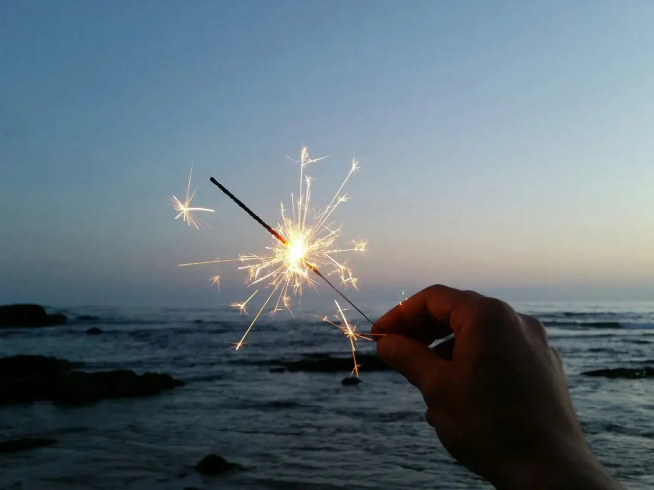Ein Wunderkerze leuchtet am Silvesterabend am Strand. Die Funken tanzen über dem ruhigen Meer und dem klaren Himmel.