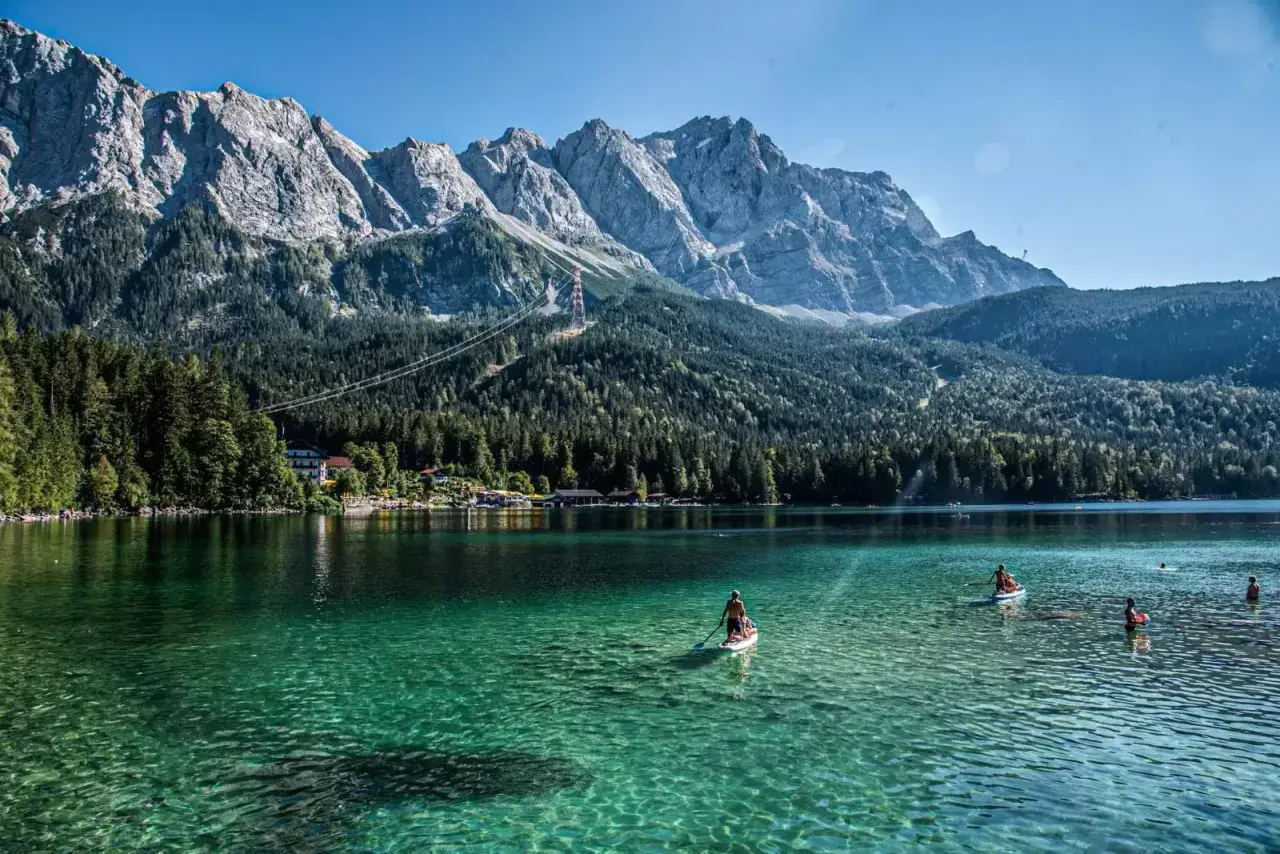 Ein Mann paddelt auf einem SUP-Board auf dem kristallklaren, blauen See in Bayern. Im Hintergrund ragen majestätische Berge auf.