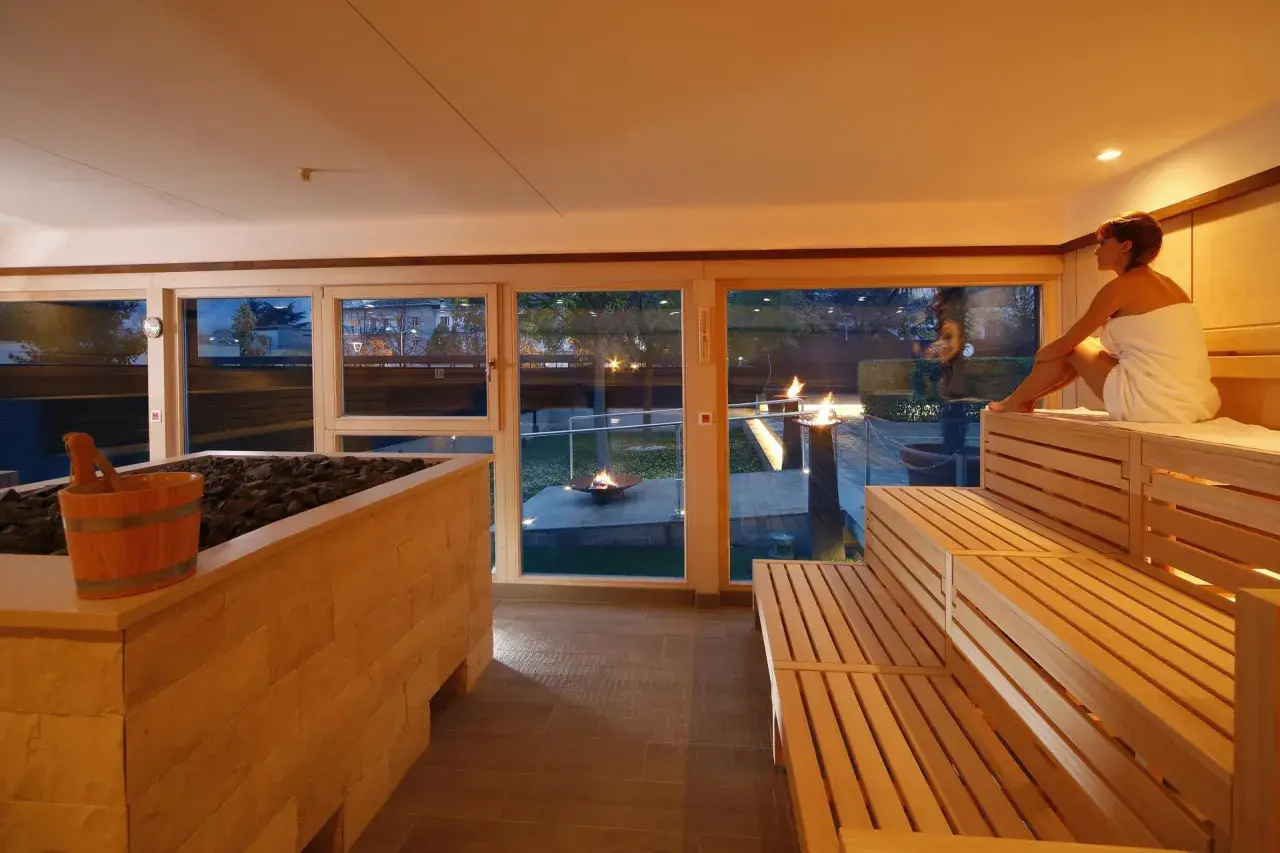 Entspannter **Sauna Bereich** mit Holzbänken und Blick auf Feuerstellen im Garten. Eine Frau in einem Handtuch sitzt auf der obersten Bank.