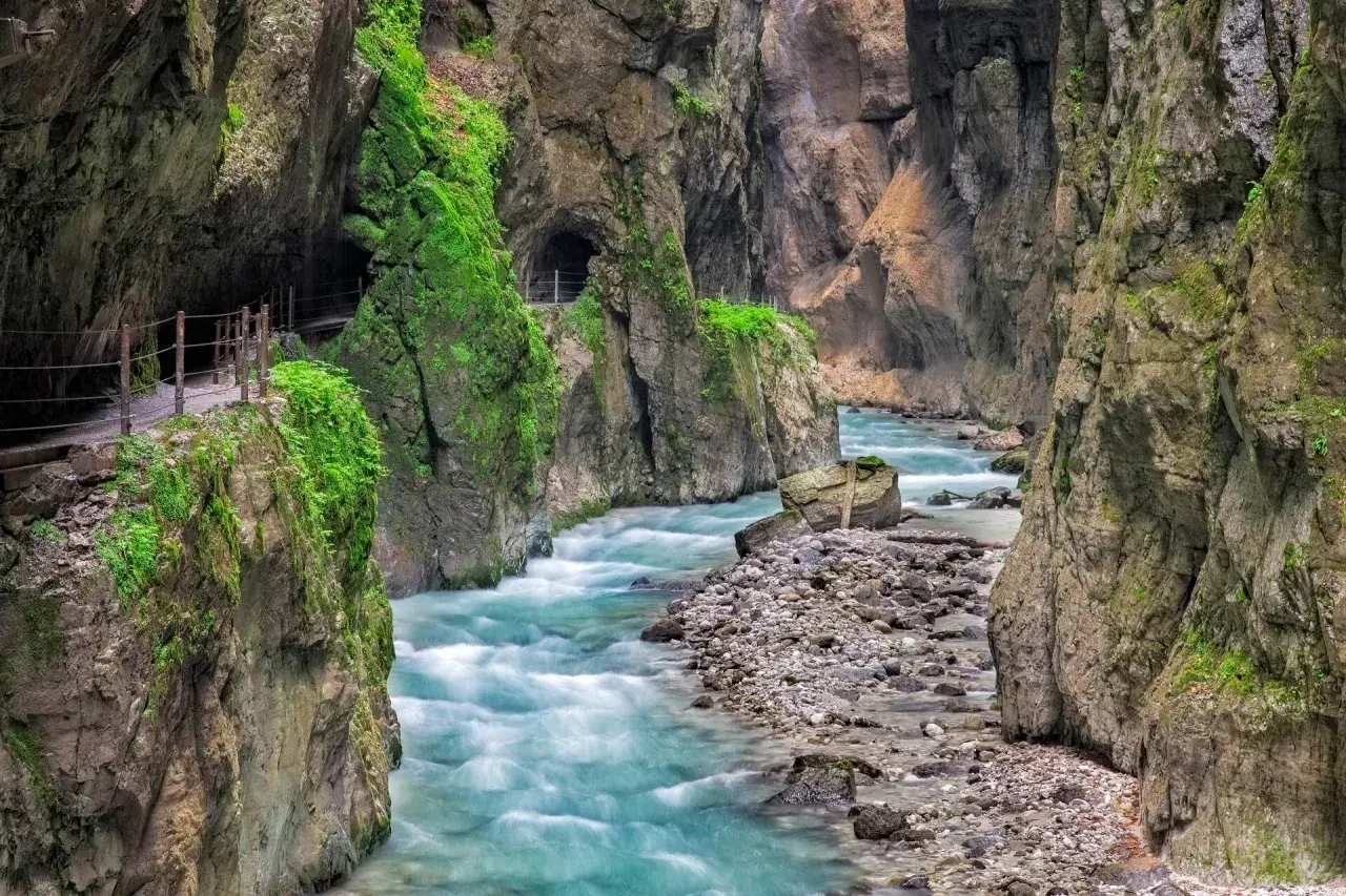 Türkisblaues Wasser rauscht durch die enge Klamm in Bayern. Ein schmaler Weg führt entlang der moosbewachsenen Felswände.