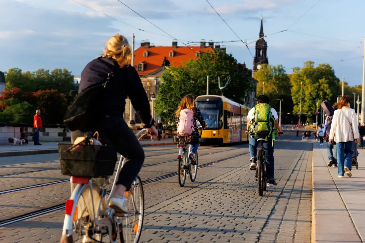 Fahrradfahrer mit Rucksäcken genießen das Bikepacking Deutschland. Eine Straßenbahn fährt vorbei, während Menschen auf Kopfsteinpflaster radeln und gehen.