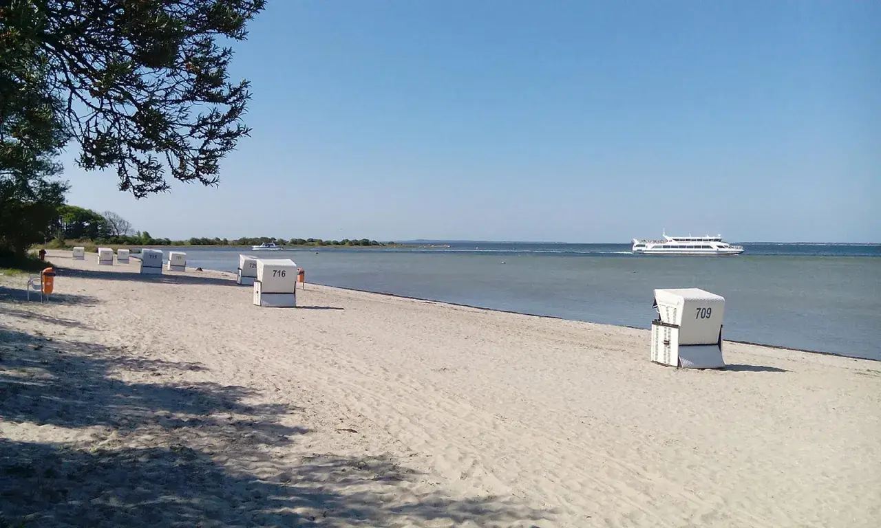 Sonniger Schaprode Strand mit Strandkörben und einem Ausflugsschiff auf dem ruhigen Meer.