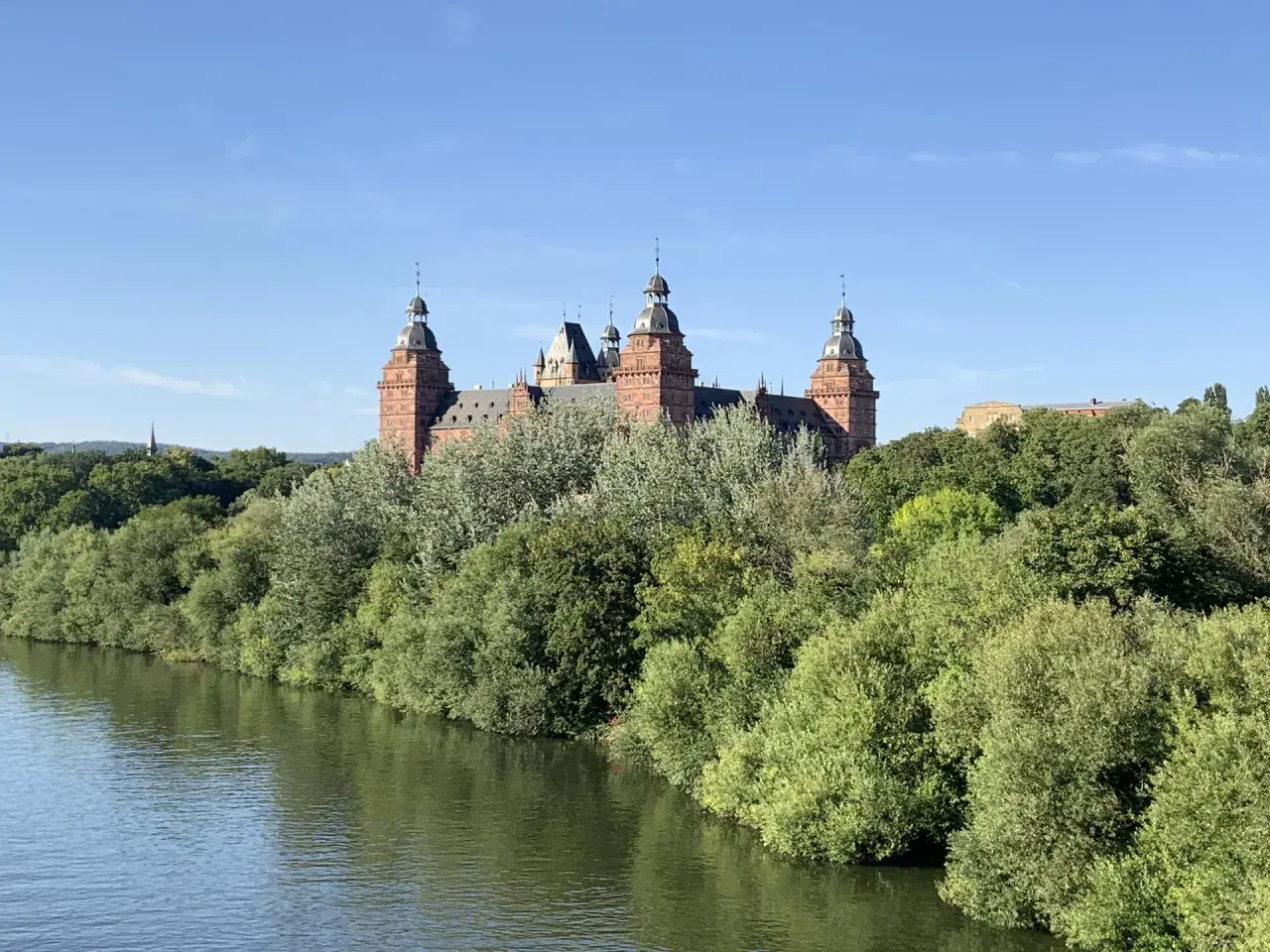 Schloss Johannisberg, ein malerisches Ziel im Spessart, ragt über üppige Bäume am Flussufer auf.