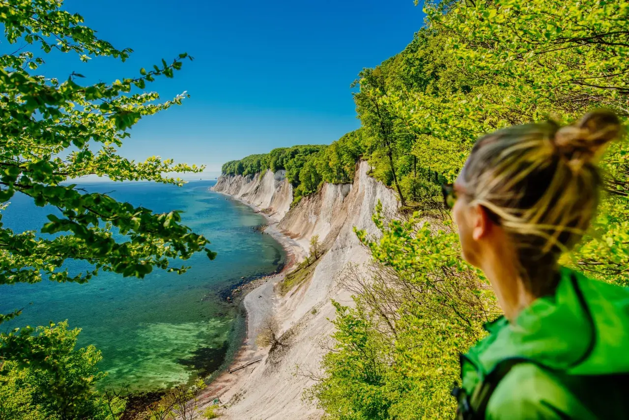 Frau blickt auf Kreidefelsen im Nationalpark Jasmund. Türkisfarbenes Meer und grüne Bäume.