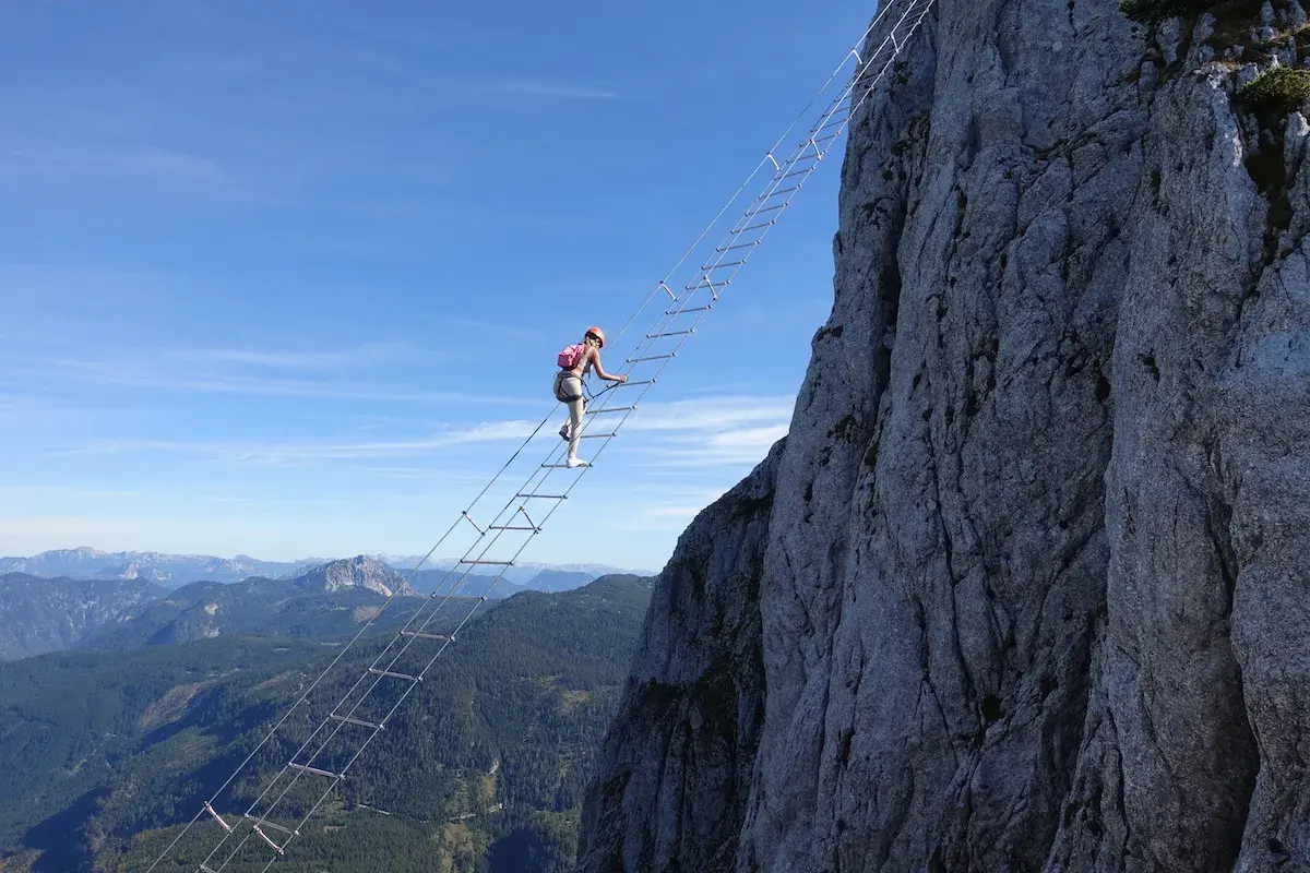 Eine Frau klettert auf einem Himmelsleiter Klettersteig eine steile Felswand empor. Die Aussicht auf die Berge ist atemberaubend.