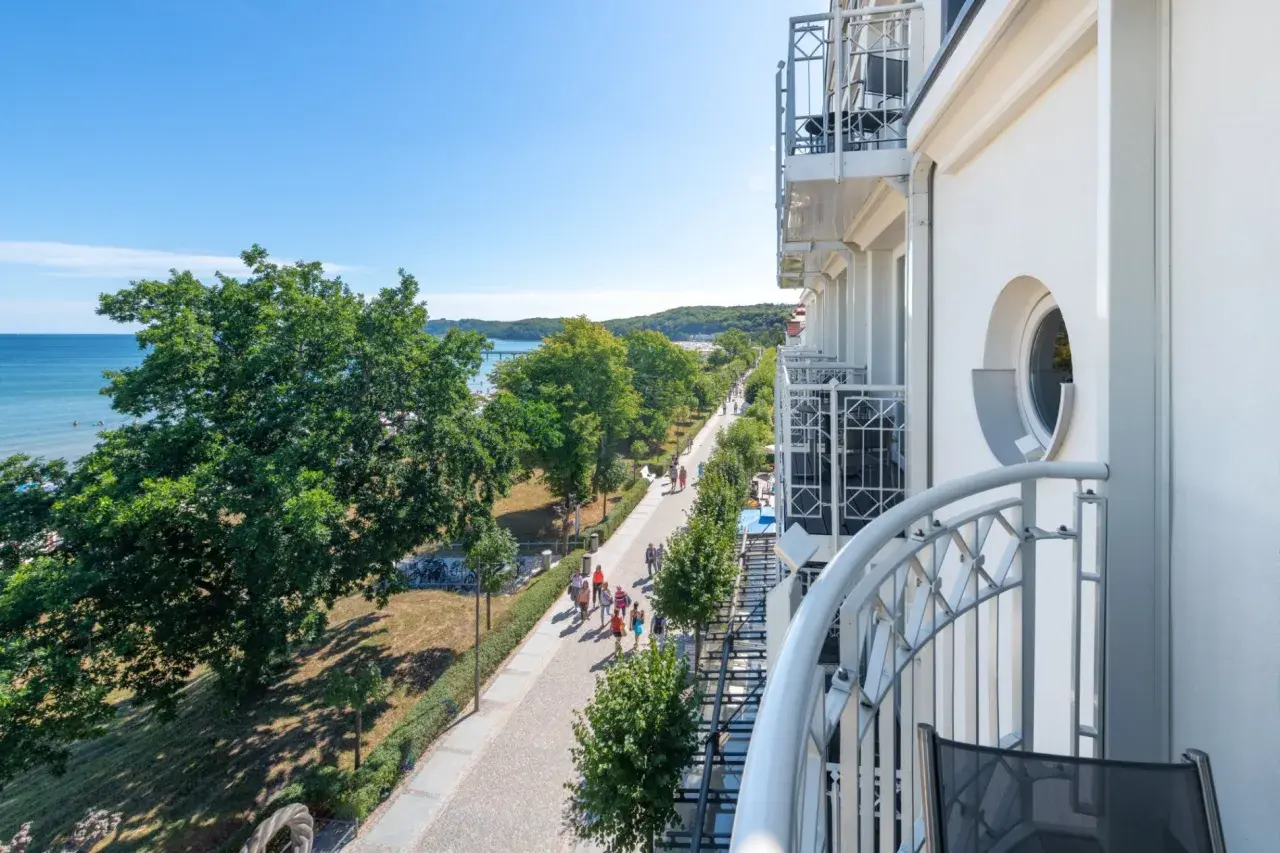 Blick von einem Balkon auf die belebte Binz Promenade mit Bäumen, Meer und Menschen, die spazieren gehen.
