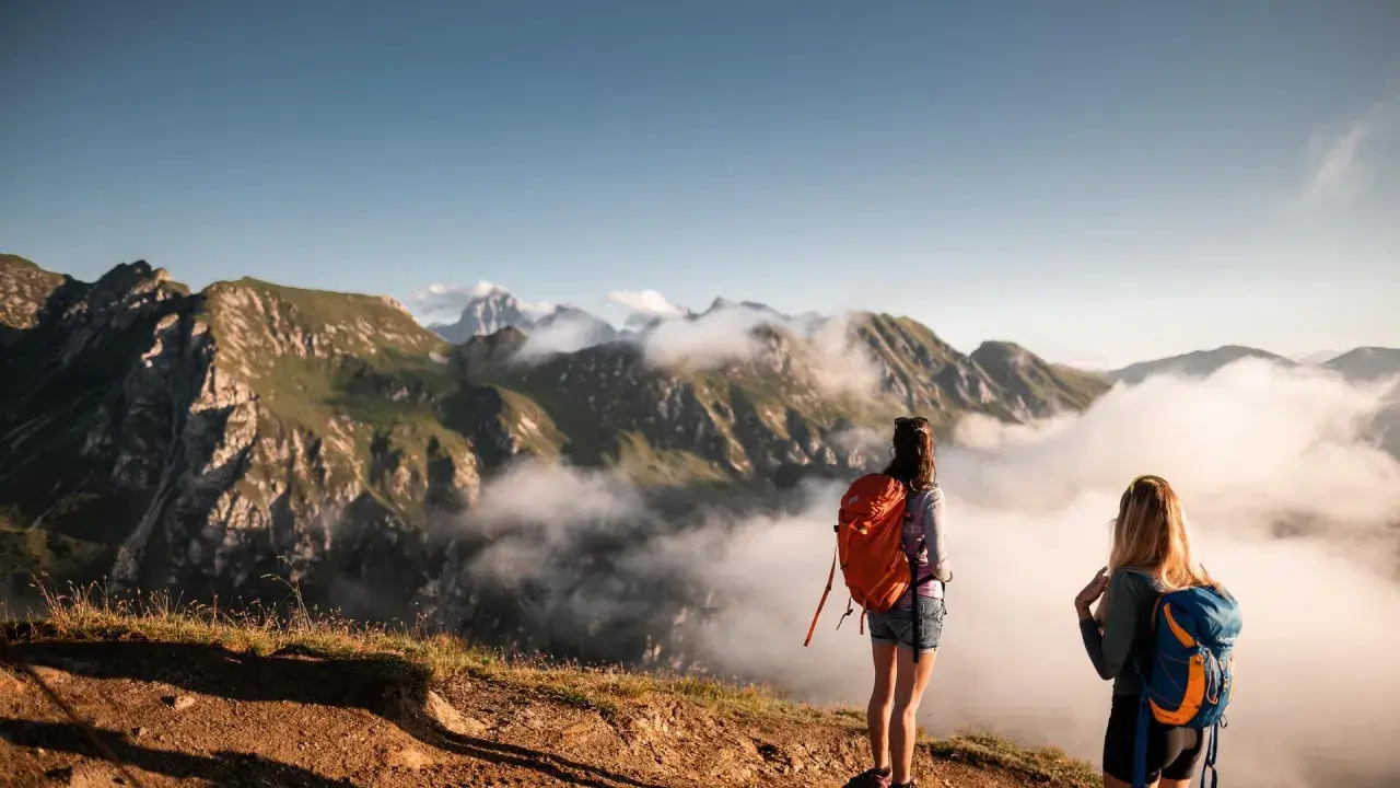 Zwei Frauen mit Rucksäcken genießen den Ausblick nach einem erfolgreichen Rosskopf Wandern. Wolken ziehen durch die Berge.