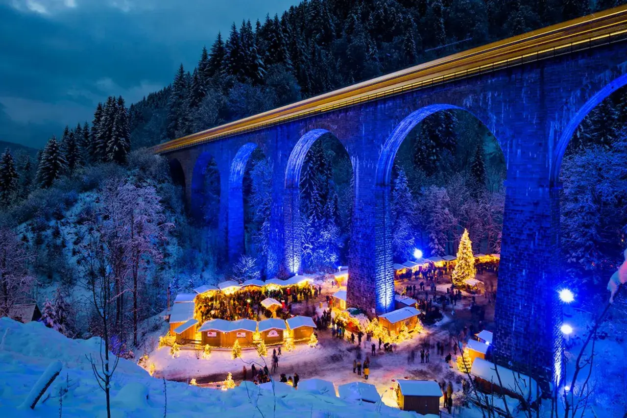 Magischer weihnachtsmarkt in deutschland unter einer beleuchteten Brücke. Ein Zug fährt vorbei, während Besucher die festliche Atmosphäre genießen.