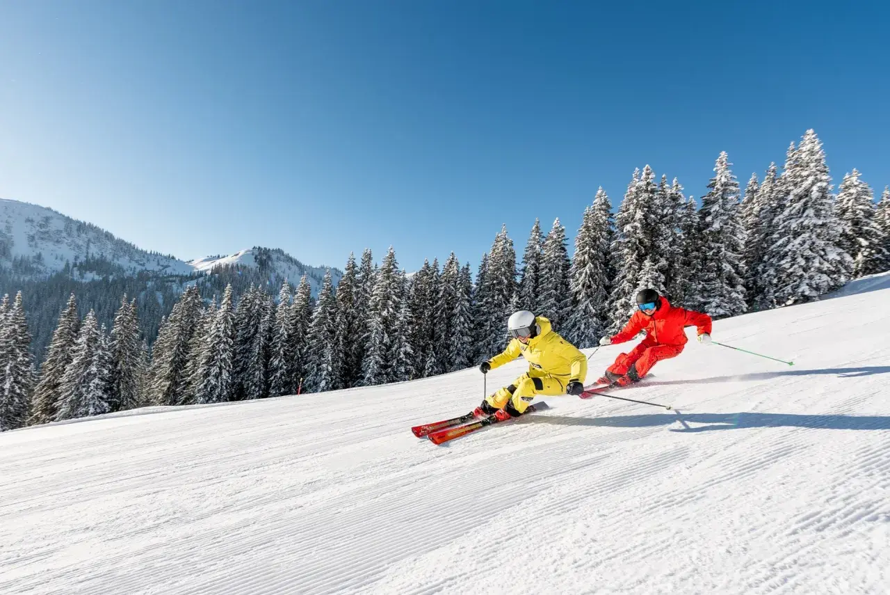 Zwei Skifahrer in leuchtenden Farben sausen über eine präparierte Piste in den **Skigebieten Alpen**. Schneebedeckte Tannen und Berge im Hintergrund.