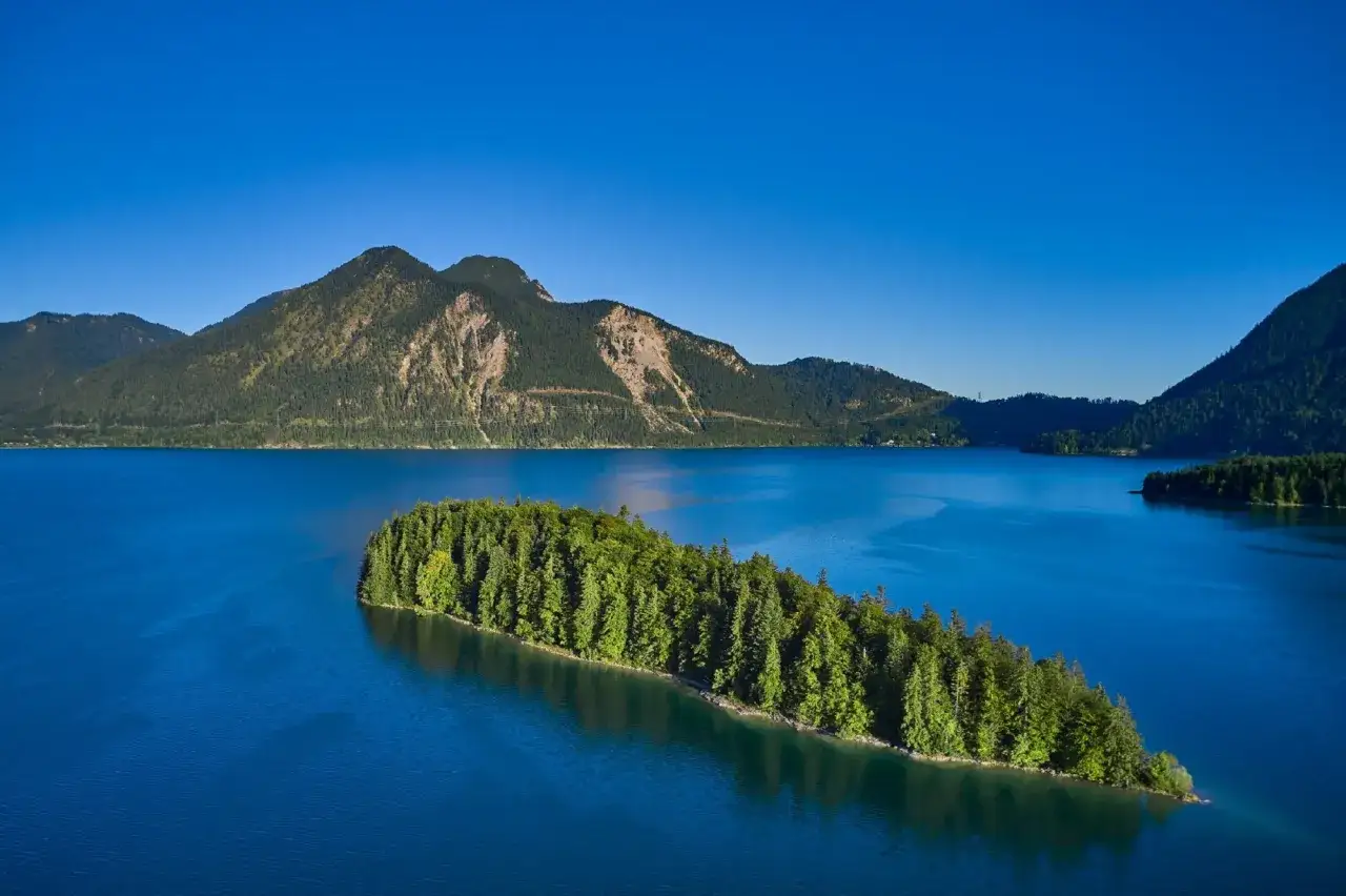 Grüne Insel mit Bäumen im tiefblauen Wasser des Walchensees in Bayern, umgeben von bewaldeten Bergen unter klarem Himmel.