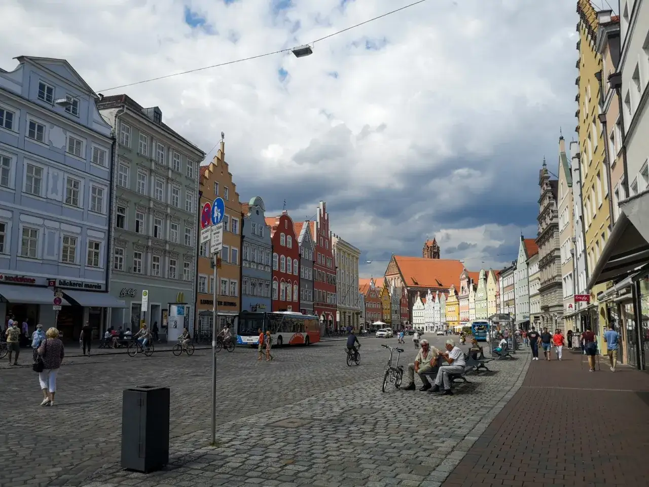 Bunte Giebelhäuser säumen den Platz, auf dem Menschen flanieren und Rad fahren. Ein Bus fährt vorbei. Entdecken Sie Landshut Geheimtipps!
