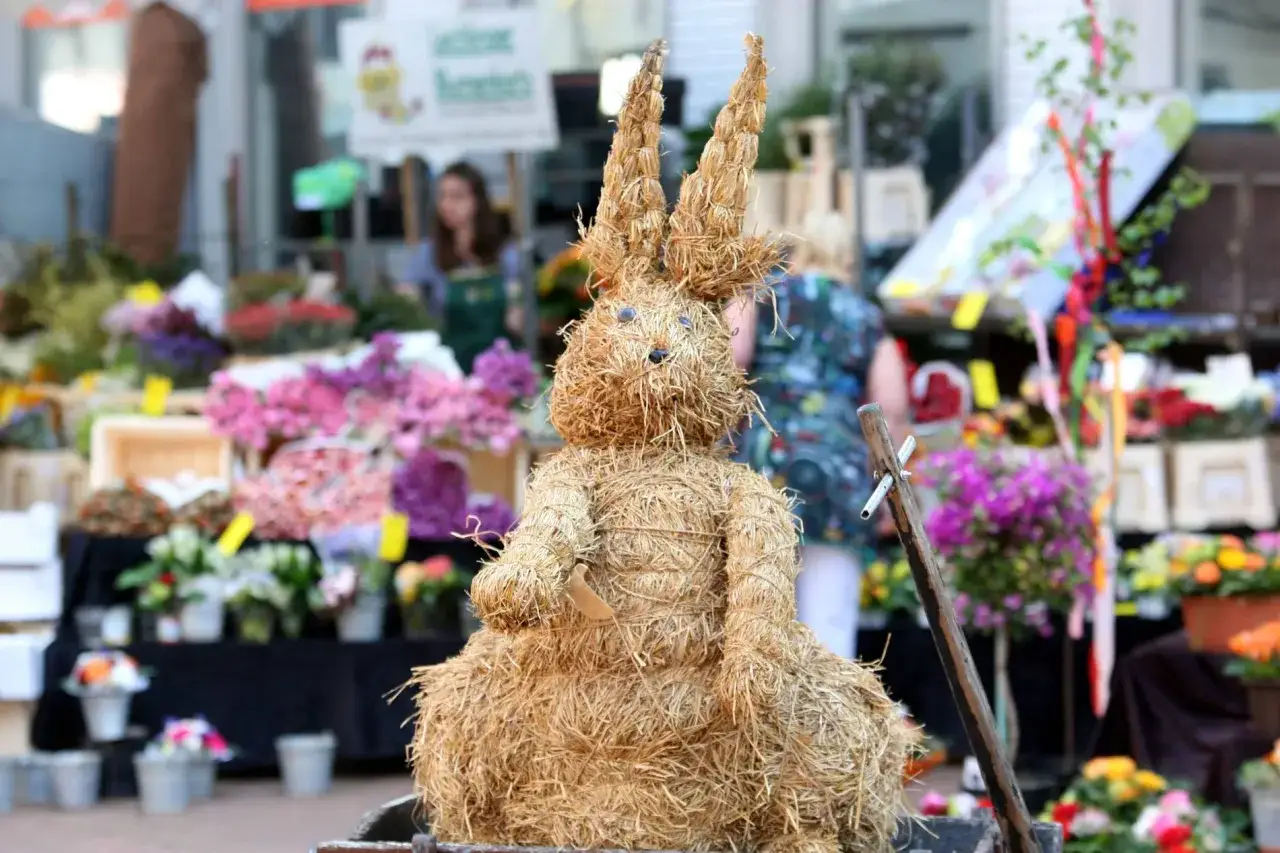 Ein Strohhase steht vor einem Blumenstand, bereit für Ostermontag-Veranstaltungen. Bunte Blumen und Dekorationen im Hintergrund.