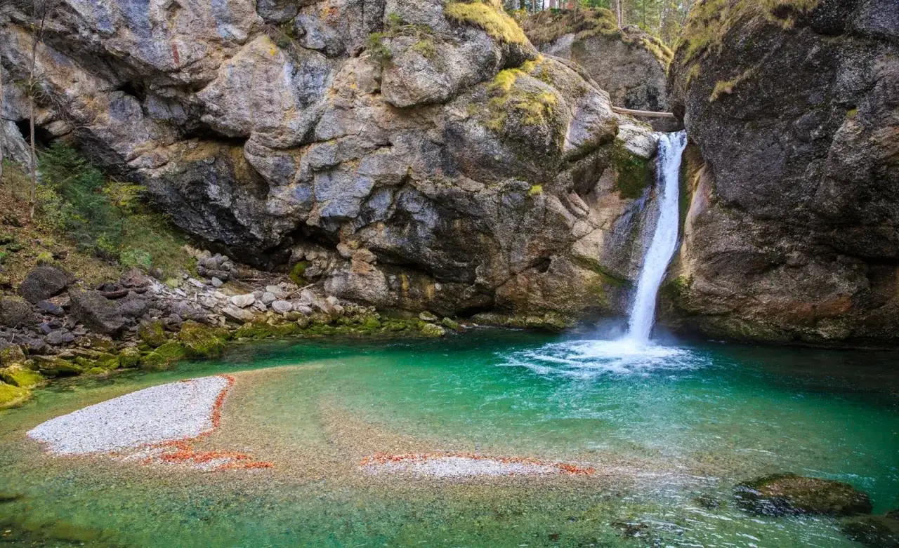 Türkiser bergsee mit einem tosenden wasserfall, der in die Tiefe stürzt. Felsen und Moos umgeben die Szenerie.