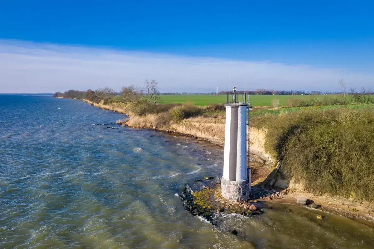 Leuchtturm auf der Halbinsel Zudar, der sich an einer felsigen Küste erhebt. Das Meer ist rau, der Himmel ist blau.