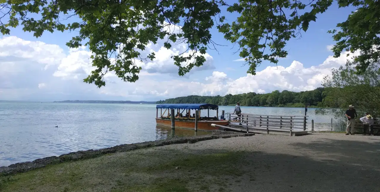 Ein Ausflugsboot wartet am Steg am Starnberger See. Im Hintergrund bewaldete Ufer und Berge.