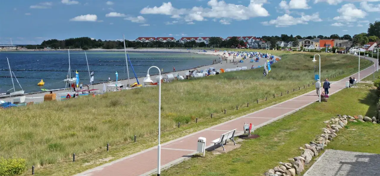 Ein sonniger Tag an der Glowe Ostsee. Boote liegen am Strand, Menschen entspannen sich unter Sonnenschirmen, und ein Weg lädt zum Spaziergang ein.
