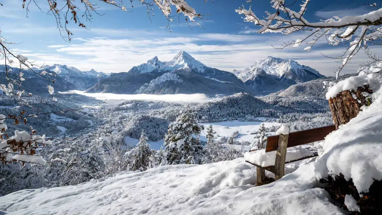 Schneebedeckte Berge und ein verschneites Tal – hier kann man im Winter Urlaub machen. Eine Bank lädt zum Verweilen ein.