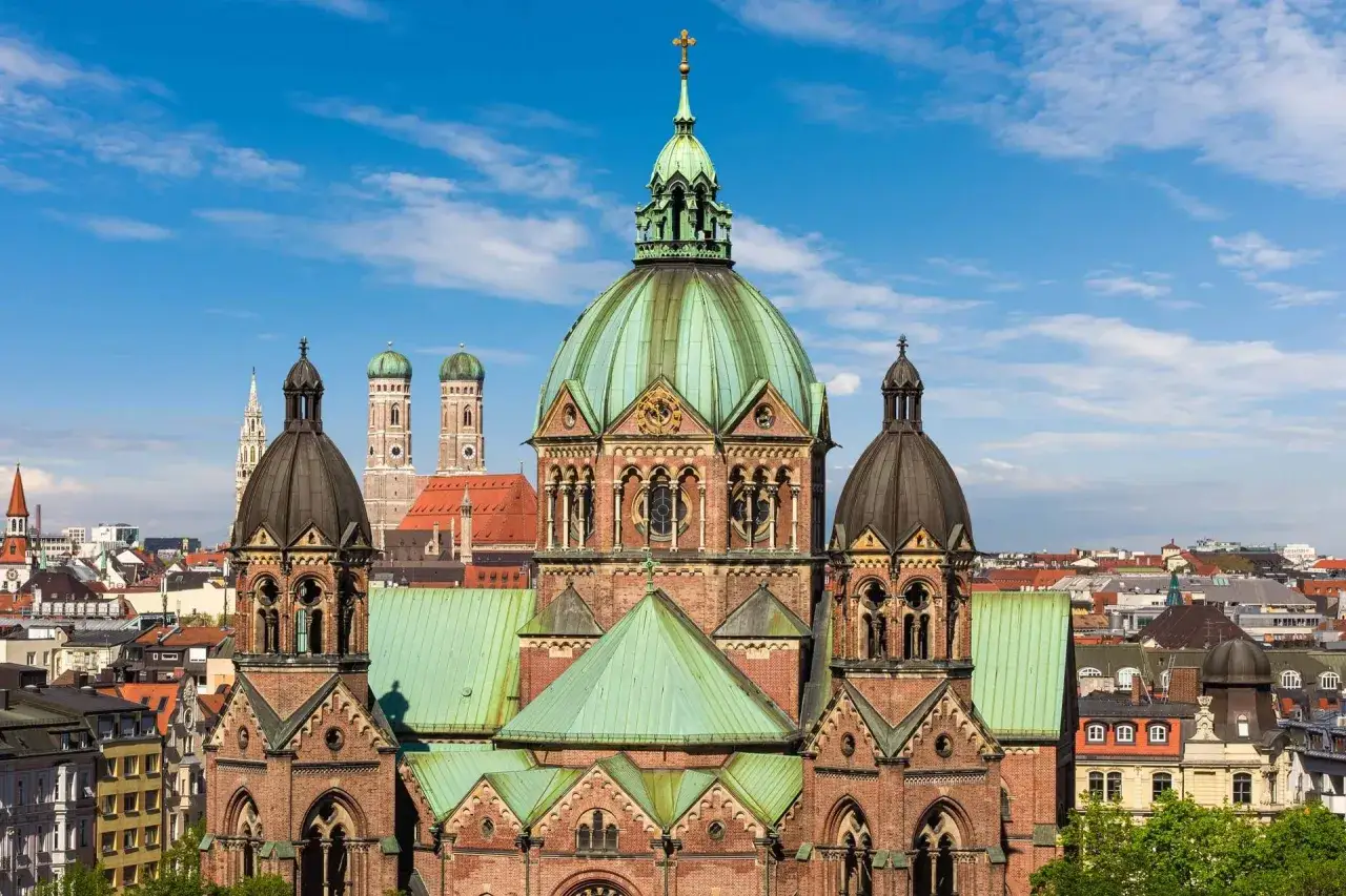 Prachtvolle Kirche mit grüner Kuppel und Türmen in Lehel, München. Im Hintergrund ragen die Türme des Münchner Doms empor.