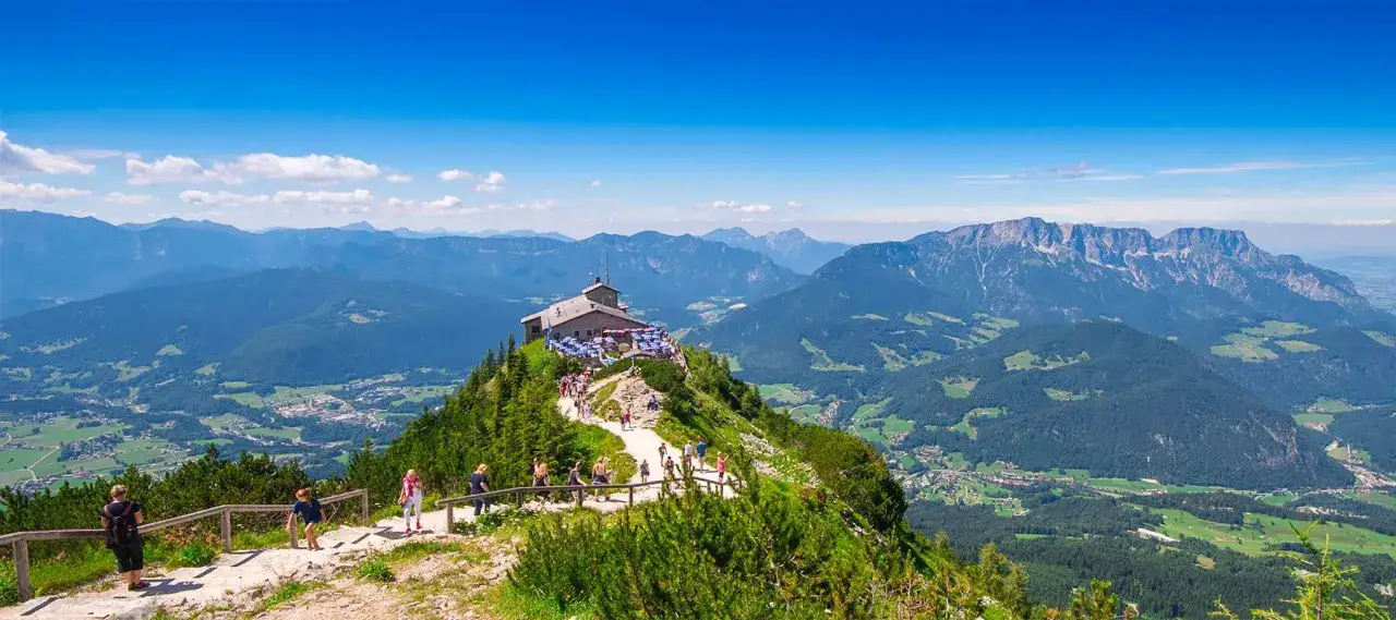 Wanderer steigen auf einem Pfad zum Kehlsteinhaus, das auf einem Berggipfel thront. Majestätische Alpenpanoramen umgeben die Szenerie.