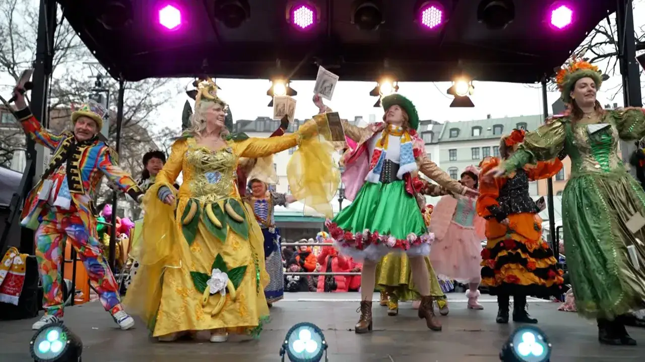 Bunte Kostüme und fröhliche Gesichter auf einer Bühne, bereit für den Fasching 2026. Ein Mann im bunten Anzug, eine Frau im gelben Kleid mit Bananen, und weitere Darsteller in fantasievollen Outfits.