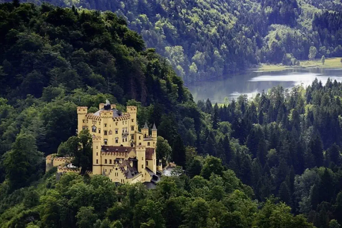 Gelbes Schloss auf einem bewaldeten Hügel, mit einem See im Hintergrund. Perfekt für Unternehmungen in Bayern.
