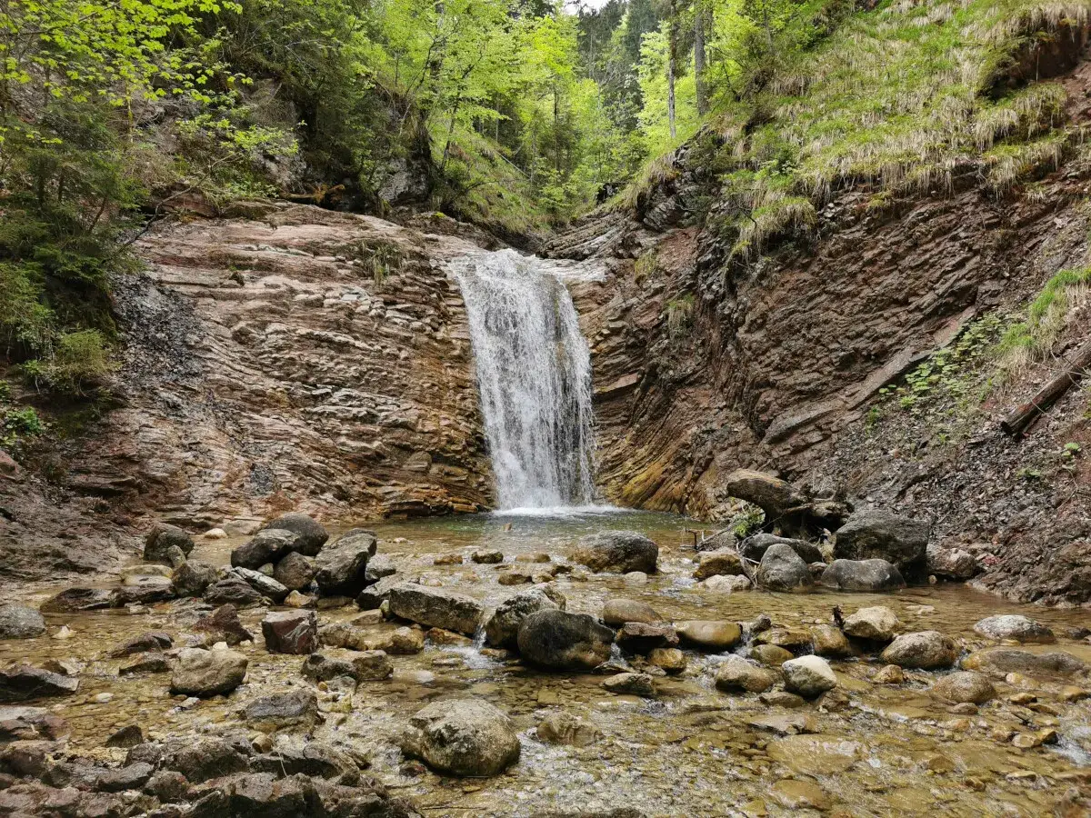 Ein malerischer Wasserfall stürzt über Felsen in ein klares Becken. Der Oberammergau Wasserfall ist umgeben von üppiger Vegetation und Felsformationen.