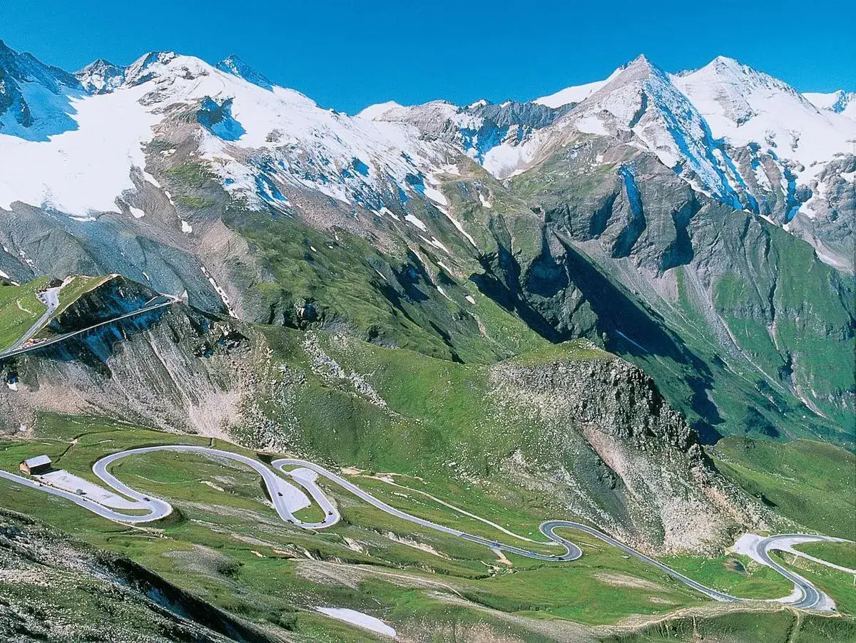 Serpentinenstra&szlig;e schl&auml;ngelt sich durch gr&uuml;ne H&auml;nge zu schneebedeckten Gipfeln. Ein perfekter Roadtrip Alpen.