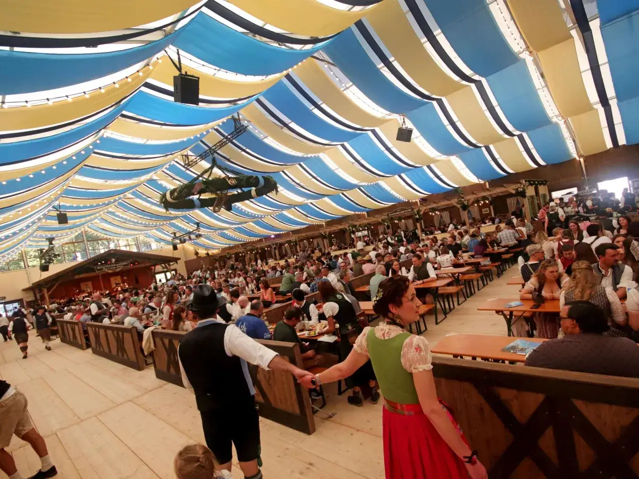 Ein Festzelt voller Menschen, die sich auf das Oktoberfest 2026 freuen. Blaue und gelbe Stoffbahnen spannen sich über das Zelt, Lichterketten leuchten.