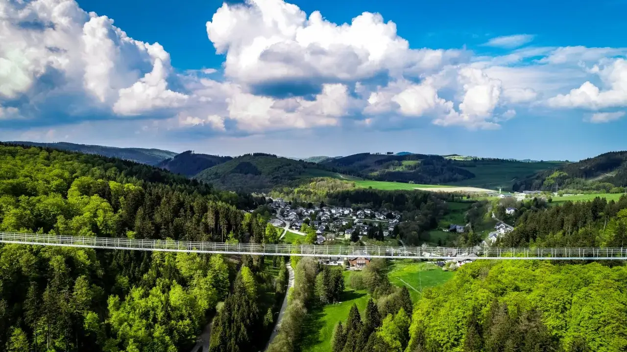 Der Skywalk Willingen überspannt ein grünes Tal mit Wäldern und einem Dorf. Wolken ziehen über den blauen Himmel.