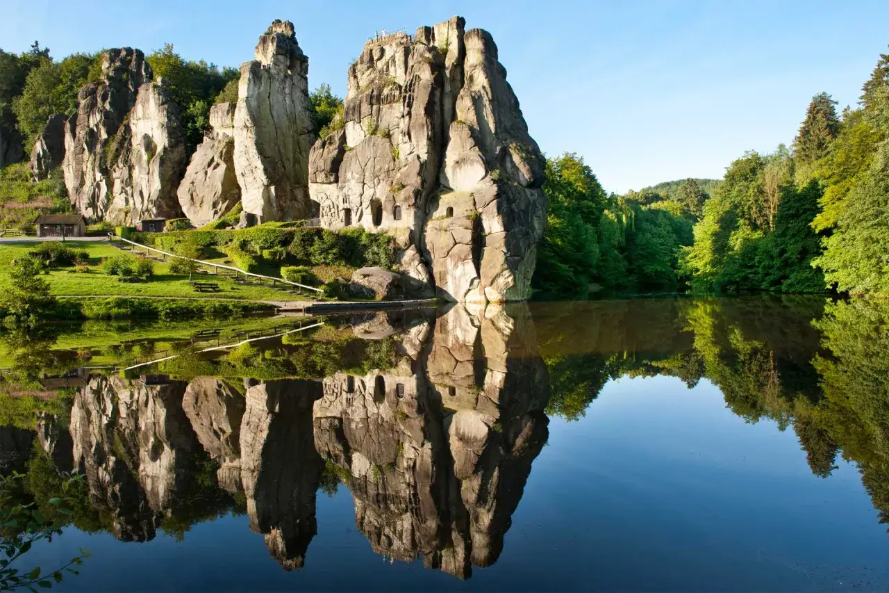 Felsformationen im Teutoburger Wald spiegeln sich in einem ruhigen See. Grüne Bäume und ein blauer Himmel umgeben die Szenerie.