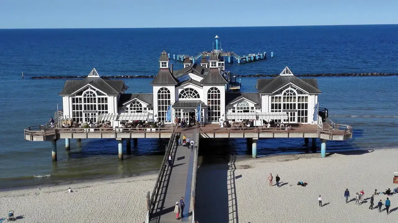 Das Kaiserpavillon-Gebäude auf der Seebrücke im Ostseebad Baabe, mit Menschen auf dem Steg und am Strand.