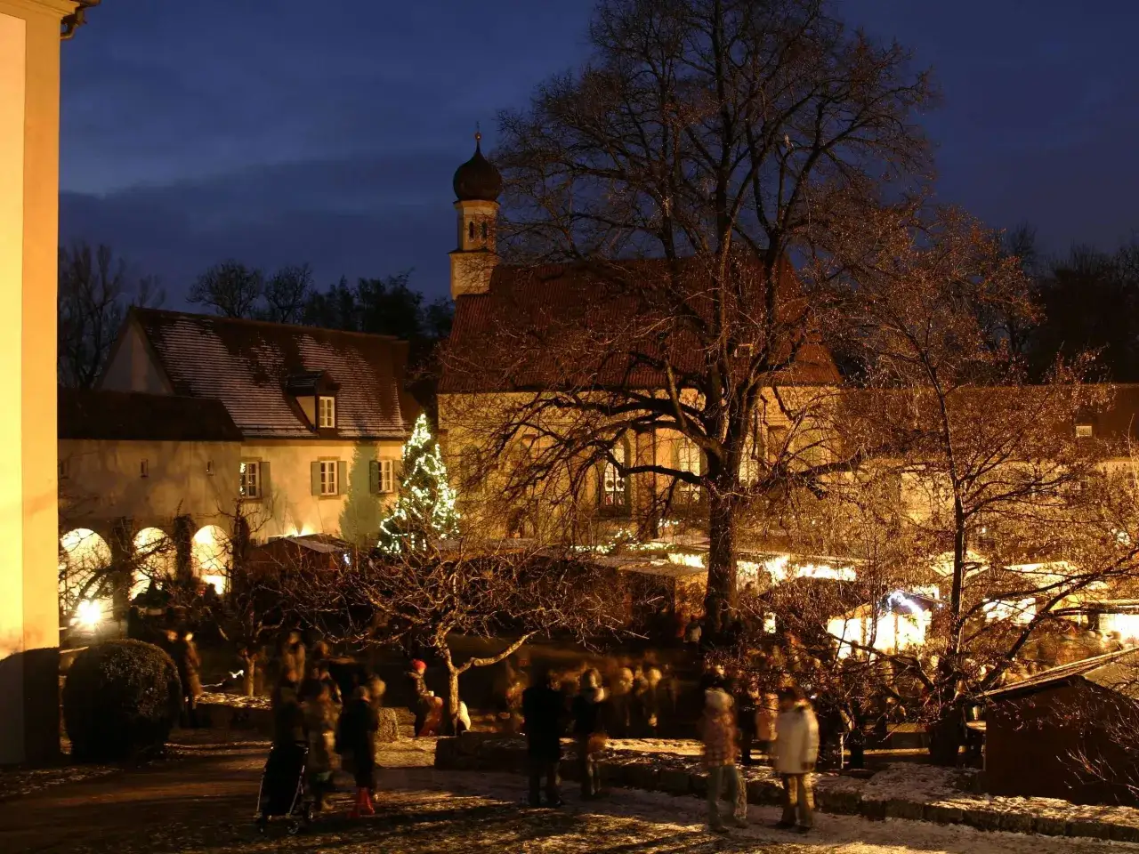 Besucher genießen den stimmungsvollen schloss nymphenburg weihnachtsmarkt im Abendlicht, mit beleuchtetem Baum und historischen Gebäuden.