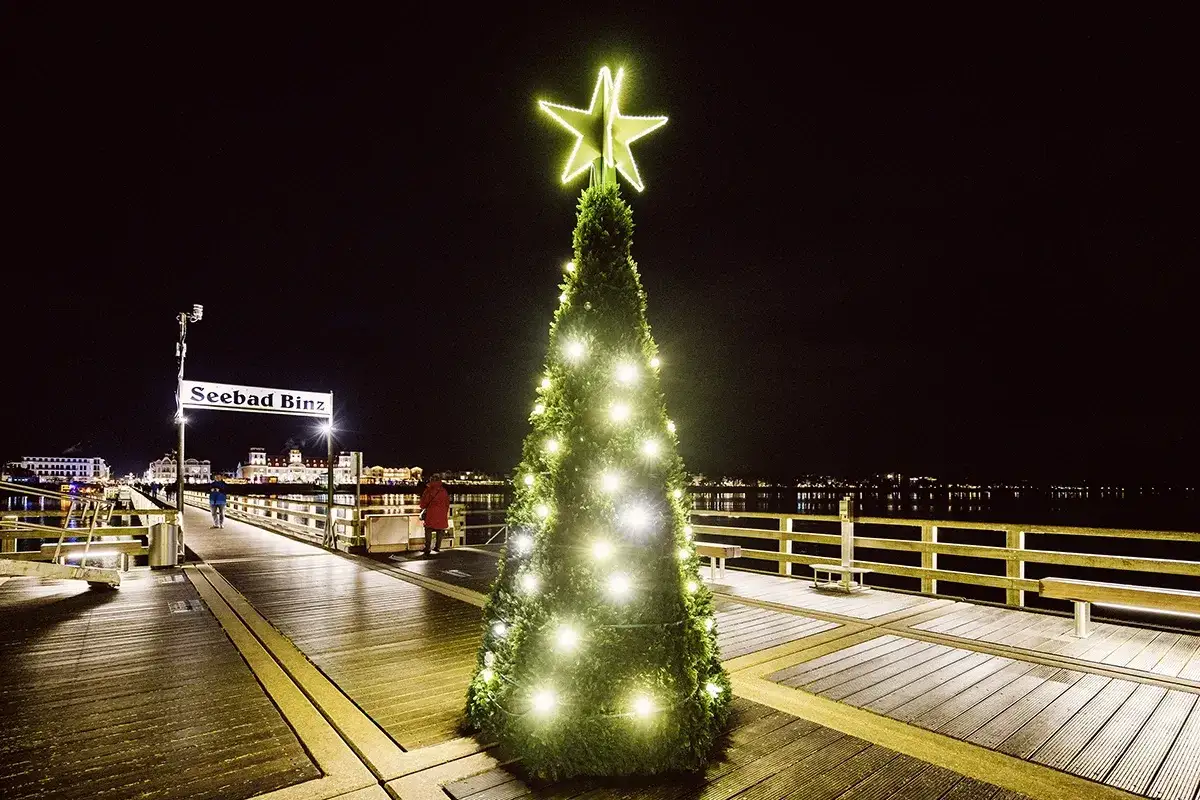 Festlich beleuchteter Weihnachtsbaum auf dem Pier in Binz. Ein Schild weist den Weg zum Seebad Binz.