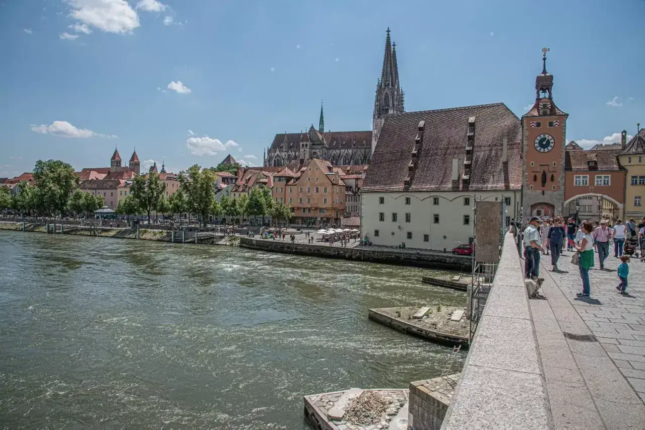 Regensburg sehenswertes: Die Donau flie&szlig;t an der historischen Altstadt vorbei, mit dem Dom St. Peter und dem Steinerernen Turm. Menschen spazieren auf der Steinernen Br&uuml;cke.