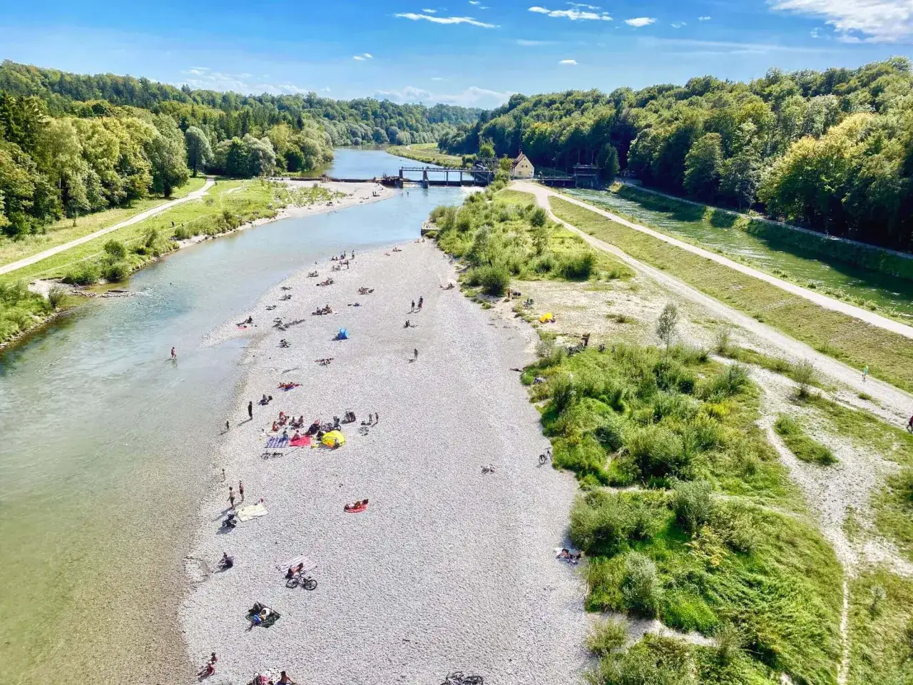 Menschen entspannen sich am Kiesstrand der Isar, während Radfahrer und Spaziergänger die Isar-Trails entlang der grünen Ufer genießen.