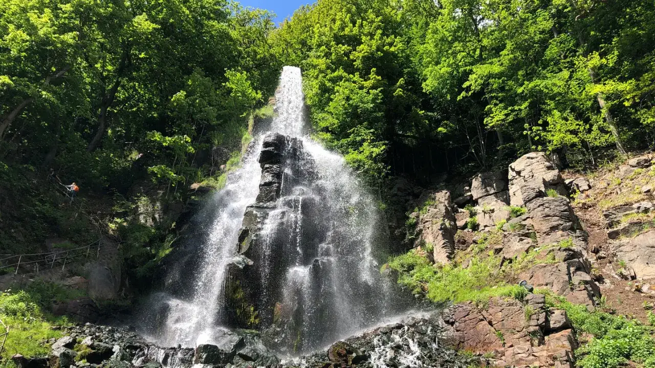 Der Trusetaler Wasserfall st&uuml;rzt &uuml;ber Felsen in die Tiefe, umgeben von &uuml;ppigem Gr&uuml;n. Ein Wanderer erklimmt den Hang.