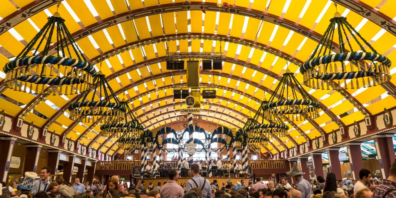 Ein festlich geschmücktes Bierzelt auf dem Oktoberfest, mit gelben Stoffbahnen unter dem Dach und großen Kronleuchtern.