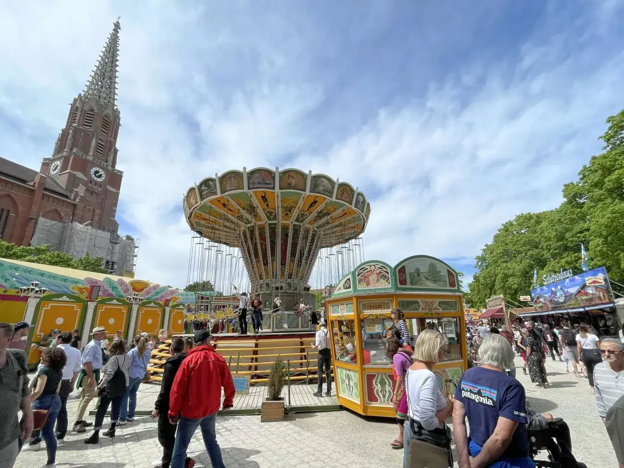 Buntes Treiben auf der Auer Dult mit einem Kettenkarussell, Buden und Menschen. Im Hintergrund ragt ein Kirchturm empor.