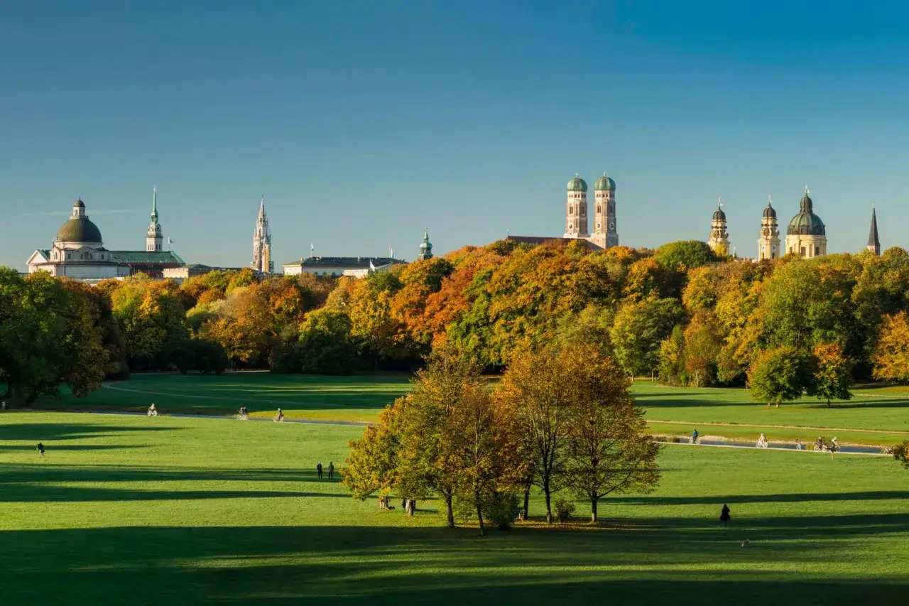 Herbstliche Farben im Englischen Garten, Schwabing, M&uuml;nchen. Menschen genie&szlig;en die Sonne auf gr&uuml;nen Wiesen, w&auml;hrend im Hintergrund die Skyline der Stadt mit ihren T&uuml;rmen und Kuppeln aufragt.