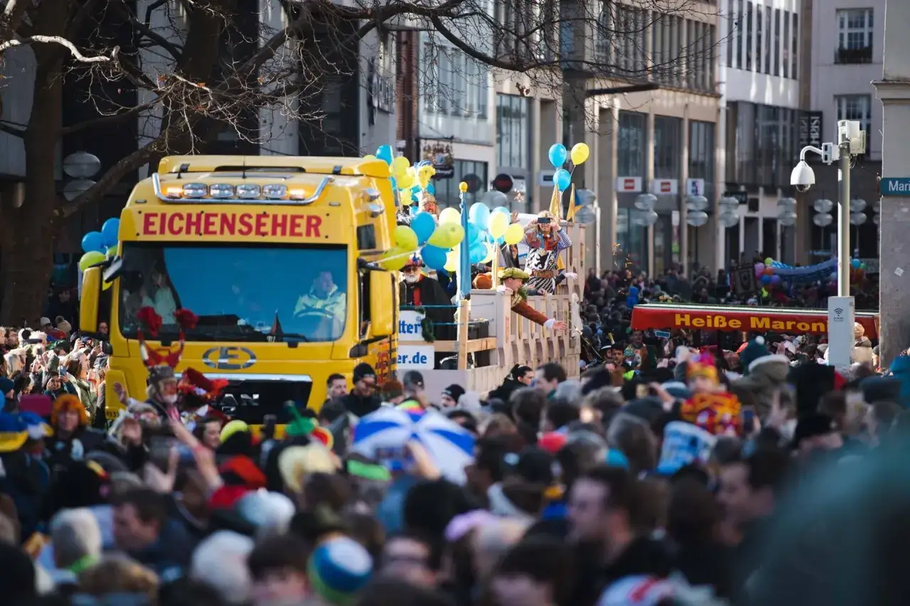 Bunte Parade beim Fasching am Marienplatz. Ein gelber LKW mit Narren und Luftballons fährt durch eine jubelnde Menge.