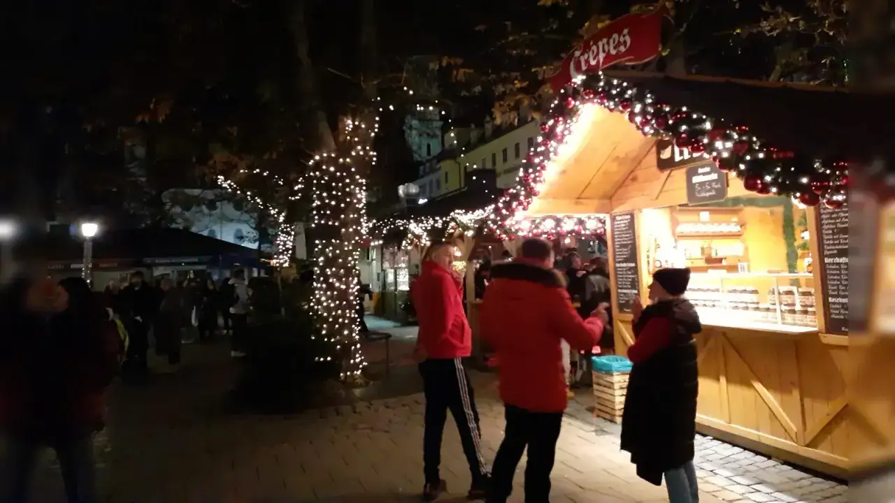 Ein festlicher winterzauber viktualienmarkt mit beleuchteten Bäumen und einem Crepes-Stand. Menschen schlendern durch die festlich geschmückte Gasse.
