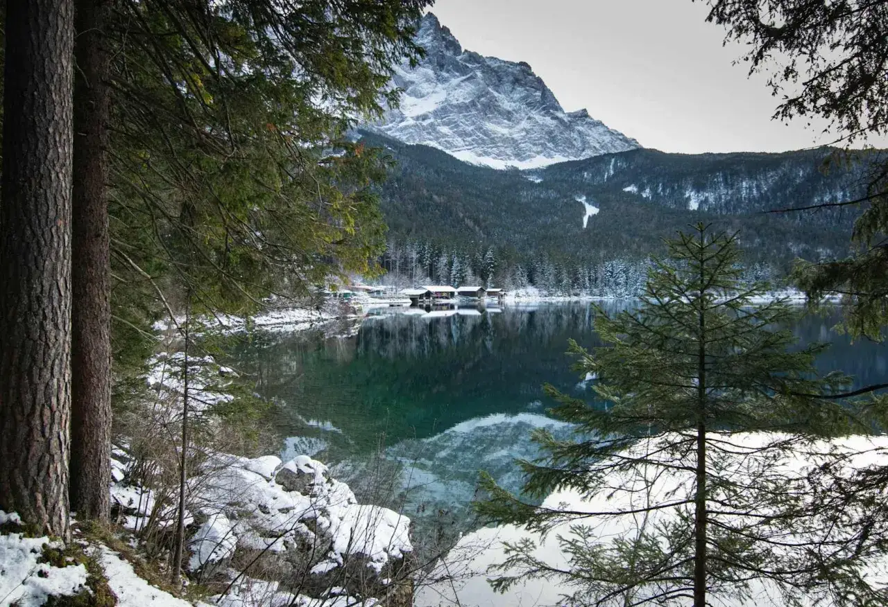 Eibsee winter: verschneite Berge spiegeln sich im klaren Wasser, H&auml;user am Ufer, B&auml;ume im Vordergrund.